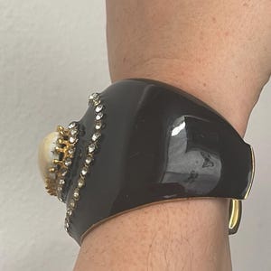 Puede incluir: Un brazalete ancho de color negro con un acento central en forma de c&uacute;pula de color crema, rodeado por un engaste dorado y un borde de peque&ntilde;os diamantes de imitaci&oacute;n transparentes. El brazalete tiene un acabado liso y brillante.