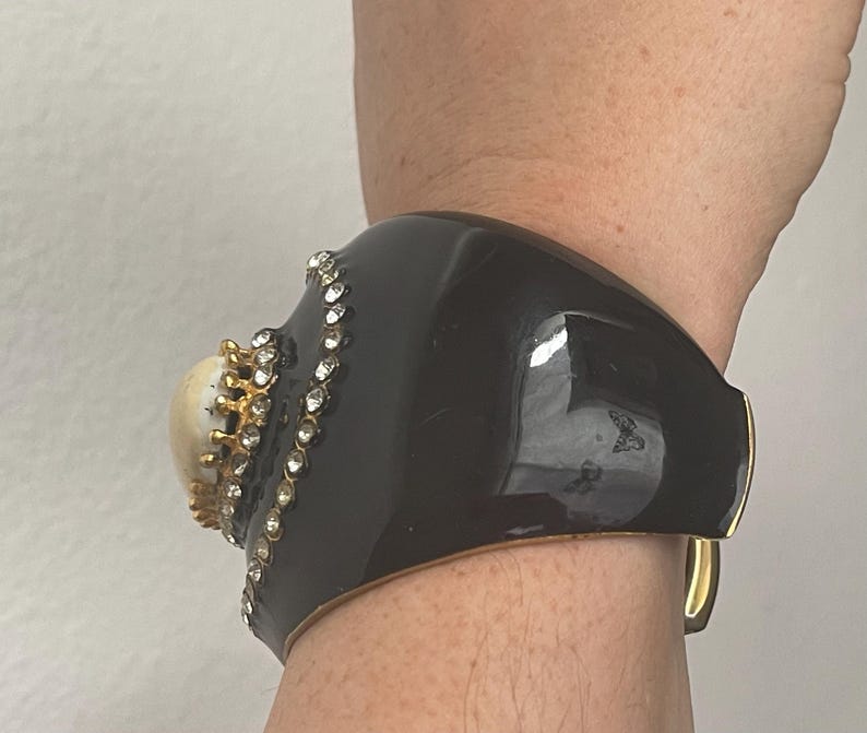Puede incluir: Un brazalete ancho de color negro con un acento central en forma de c&uacute;pula de color crema, rodeado por un engaste dorado y un borde de peque&ntilde;os diamantes de imitaci&oacute;n transparentes. El brazalete tiene un acabado liso y brillante.
