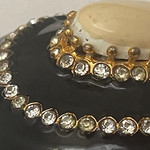 Puede incluir: Un objeto redondeado de color negro brillante con dos bandas met&aacute;licas doradas conc&eacute;ntricas incrustadas con brillantes transparentes. Una piedra circular opaca de color crema est&aacute; situada en el centro, rodeada por un borde decorativo de metal dorado.