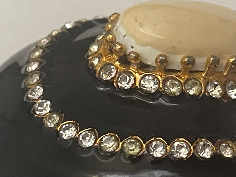 Puede incluir: Un objeto redondeado de color negro brillante con dos bandas met&aacute;licas doradas conc&eacute;ntricas incrustadas con brillantes transparentes. Una piedra circular opaca de color crema est&aacute; situada en el centro, rodeada por un borde decorativo de metal dorado.