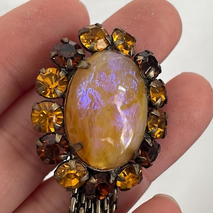May include: A vintage brooch with an oval, iridescent, yellow-orange stone, encircled by amber and brown rhinestones. It has a metal chain fringe. The brooch is held in a hand.
