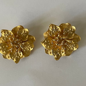 May include: A pair of gold-tone flower-shaped earrings. The earrings are made of metal and have a textured surface.