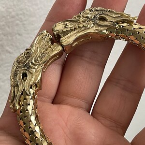 May include: Gold-toned bracelet featuring dragon head clasps. The bracelet's design includes intricate dragon head details and a flexible chain-mail style band. The bracelet is a striking piece of jewelry.