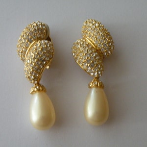 May include: A pair of gold-toned clip-on earrings. The upper portion is encrusted with small, clear crystals. A teardrop-shaped, cream-colored pearl dangles from each earring, creating an elegant and classic look. The earrings are set against a plain white background.