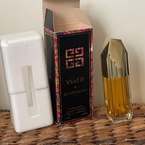 May include: A bottle of Ysatis de Givenchy perfume with a gold cap and a clear glass body, next to its box and a white foam insert. The box is black with a colorful floral design and the brand name.