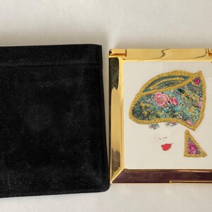 May include: A gold-framed compact mirror with an illustration of a woman wearing a floral hat. The compact is next to a black velvet pouch. The woman's hat has pink roses and green leaves.