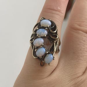 May include: A vintage ring with a gold-toned metal setting. The ring features five oval, light blue opals arranged vertically. The setting has an ornate, swirling design around the opals, creating an elegant and classic look.