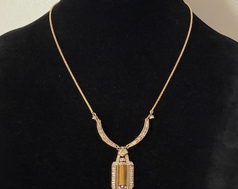 May include: Gold-toned necklace with a rectangular pendant featuring a tiger's eye stone. The pendant is framed with small, sparkling accents. The necklace is displayed on a black mannequin, showcasing its design and details.