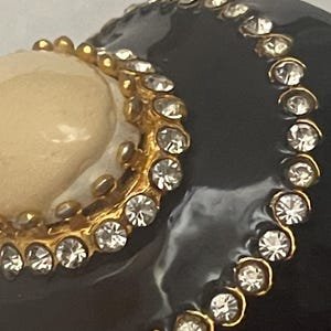 Puede incluir: Un objeto curvo de color negro brillante con un acento circular liso de color crema bordeado por un marco de metal dorado engastado con peque&ntilde;os diamantes de imitaci&oacute;n transparentes. Una segunda l&iacute;nea m&aacute;s larga de brillantes sigue la curva.