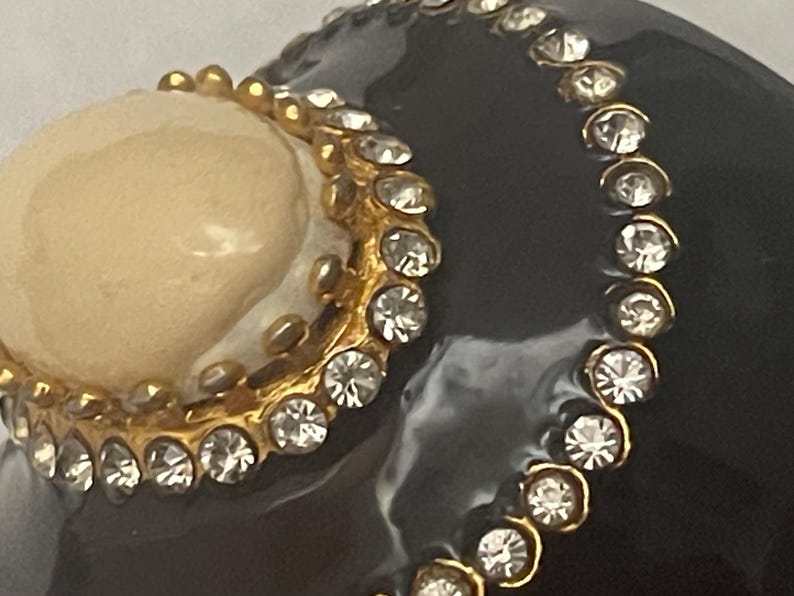 Puede incluir: Un objeto curvo de color negro brillante con un acento circular liso de color crema bordeado por un marco de metal dorado engastado con peque&ntilde;os diamantes de imitaci&oacute;n transparentes. Una segunda l&iacute;nea m&aacute;s larga de brillantes sigue la curva.
