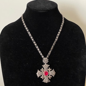 May include: A silver necklace with a large, ornate cross pendant featuring a red gemstone. The chain is made of small, linked flower shapes.
