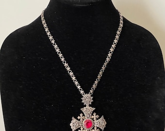 May include: A silver necklace with a large, ornate cross pendant featuring a red gemstone. The chain is made of small, linked flower shapes.