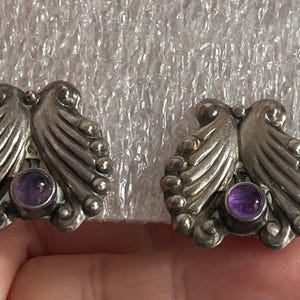 以下が含まれることがあります： 銀色のスタッドピアス一組。各イヤリングは、中央に丸い紫のアメジストストーンが付いた、様式化された翼のデザインが特徴です。イヤリングは、テクスチャ加工された白い背景に配置されています。