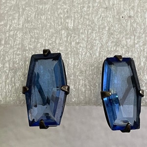 Könnte beinhalten: Ein Paar rechteckige blaue Edelstein-Ohrstecker. Die Ohrringe haben eine dunkle Metalleinfassung mit kleinen Akzenten oben und unten. Die Edelsteine sind leuchtend blau und facettiert.
