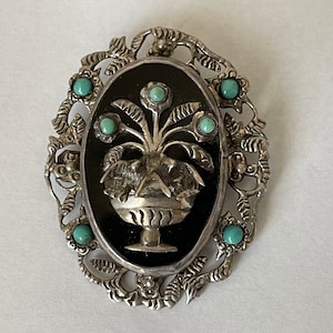 May include: A silver brooch with a black oval cameo featuring a silver floral design with turquoise stones. The cameo is surrounded by a silver filigree frame with turquoise stones.