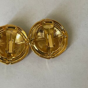 May include: A pair of gold-tone clip-on earrings with a circular design. The earrings have a textured surface and a simple, elegant style.