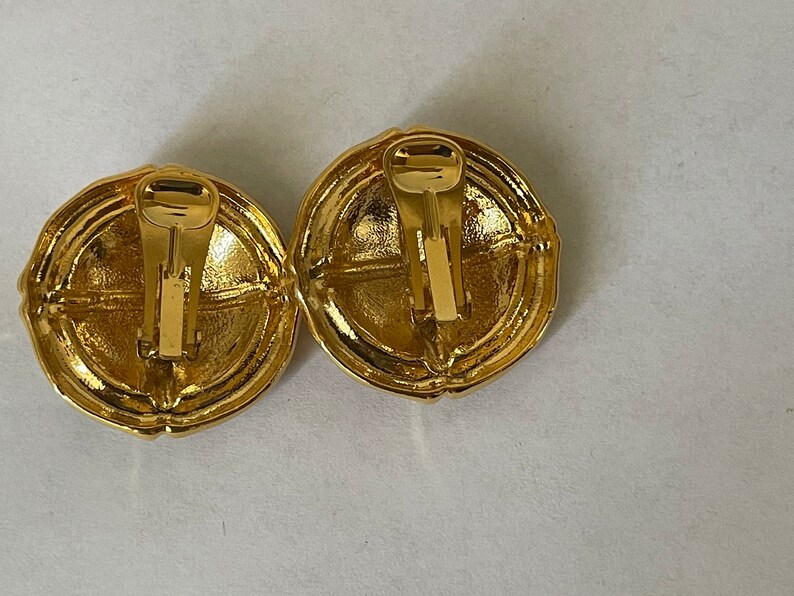 May include: A pair of gold-tone clip-on earrings with a circular design. The earrings have a textured surface and a simple, elegant style.