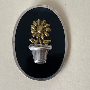 Puede incluir: Un broche de plata y oro con un girasol estilizado en una maceta. El girasol es de oro y la maceta es de plata. El broche está sobre un fondo negro.