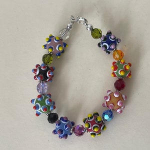 Puede incluir: Un brazalete colorido con un cierre plateado, que presenta una variedad de cuentas de vidrio con patrones en tonos de azul, verde, rojo, amarillo y rosa.