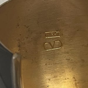 Puede incluir: Un brazalete dorado con una superficie interior lisa y pulida. La banda interior presenta el texto grabado "MIGHT" colocado sobre un logotipo "V" estilizado encerrado dentro de un contorno rectangular, mostrando detalles met&aacute;licos claros.
