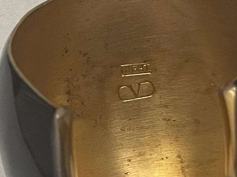 Puede incluir: Un brazalete dorado con una superficie interior lisa y pulida. La banda interior presenta el texto grabado "MIGHT" colocado sobre un logotipo "V" estilizado encerrado dentro de un contorno rectangular, mostrando detalles met&aacute;licos claros.