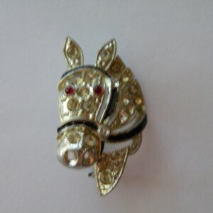 Pode incluir: Um broche de cabeça de cavalo prateado com olhos de strass vermelhos e detalhes de strass transparentes.