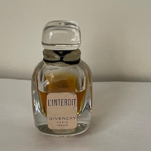 Puede incluir: Un pequeño frasco de perfume de vidrio transparente, de forma cuadrada y con tapón transparente. El frasco contiene un líquido de color dorado y tiene la etiqueta "L'INTERDIT GIVENCHY PARIS FRANCE."