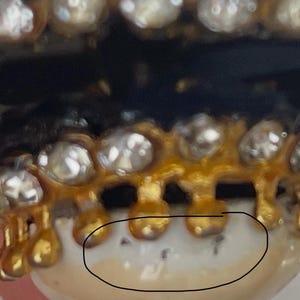 Puede incluir: Un grill dental dorado con una hilera de piedras transparentes de corte redondo a lo largo del borde superior. La estructura met&aacute;lica se extiende hacia abajo en peque&ntilde;as puntas redondeadas sobre una superficie blanca similar a un diente.