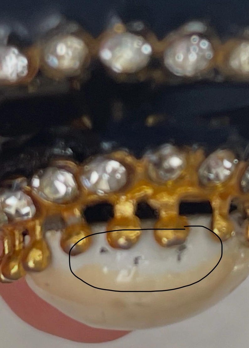 Puede incluir: Un grill dental dorado con una hilera de piedras transparentes de corte redondo a lo largo del borde superior. La estructura met&aacute;lica se extiende hacia abajo en peque&ntilde;as puntas redondeadas sobre una superficie blanca similar a un diente.
