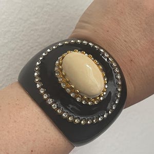 Puede incluir: Una pulsera brazalete ancha de color negro con una piedra cabuj&oacute;n ovalada de color crema en el centro, engastada en un marco de metal dorado. La piedra est&aacute; rodeada por un borde circular de peque&ntilde;os brillantes transparentes en la superficie.