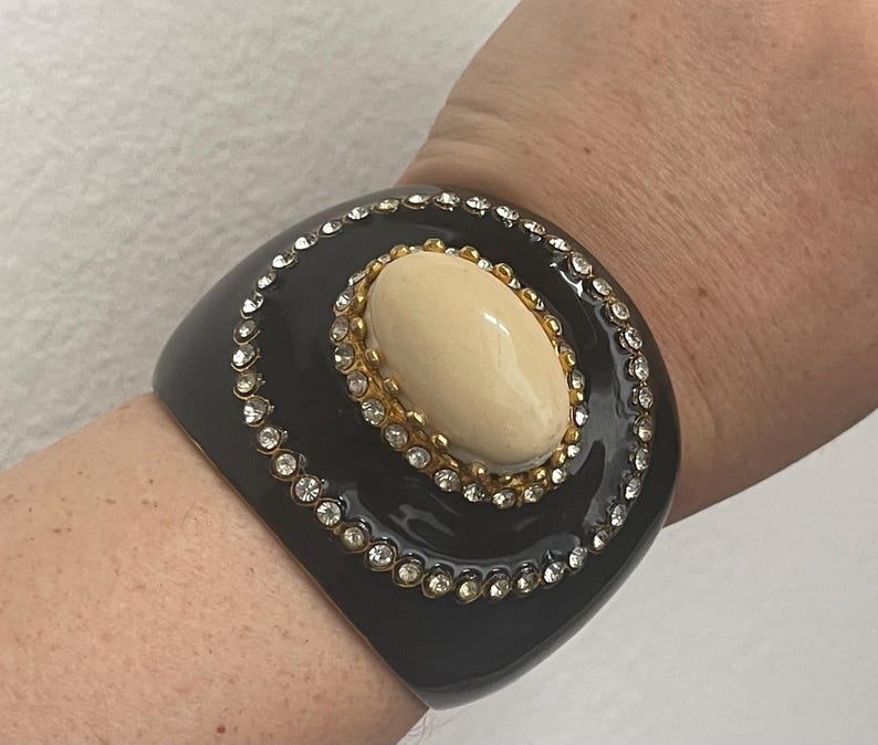 Puede incluir: Una pulsera brazalete ancha de color negro con una piedra cabuj&oacute;n ovalada de color crema en el centro, engastada en un marco de metal dorado. La piedra est&aacute; rodeada por un borde circular de peque&ntilde;os brillantes transparentes en la superficie.