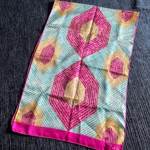 May include: A white silk scarf with a geometric pattern of pink, yellow, and blue hexagons. The scarf has a pink border and the word "Echo" is printed in the bottom right corner.