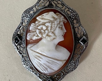May include: A cameo brooch with a carved shell cameo of a woman's profile, set in a silver filigree frame with two black gemstones.