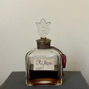 May include: A vintage perfume bottle with a clear glass body and a tulip-shaped stopper. The bottle contains a dark amber liquid and is labeled "Réplique, Raphael Paris". A red wax seal hangs from the neck.