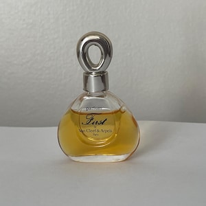 May include: A clear glass perfume bottle with a golden liquid inside, featuring a silver-toned circular cap. The front of the bottle displays the text "parfum First de Van Cleef & Arpels Paris" in black lettering on the glass surface.