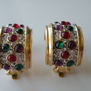 May include: A pair of gold tone hoop earrings with a row of red, green, and purple gemstones set in a gold tone metal frame. The earrings have a clip-on closure.