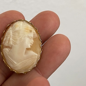 May include: A gold-toned cameo brooch featuring a carved shell cameo of a woman's profile. The cameo is set in a gold-toned metal frame with a scalloped edge.