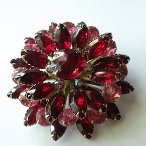 Könnte beinhalten: Eine Brosche in Form einer Blume aus roten und rosafarbenen Strasssteinen. Die Brosche ist aus Metall und hat eine goldene Oberfläche.