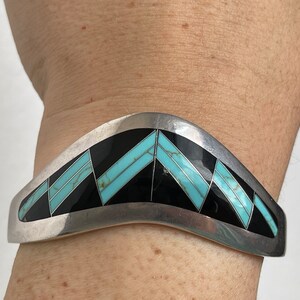 May include: A silver bracelet with a geometric design featuring turquoise and black stones.