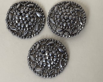 May include: Three decorative, dark gray, round buttons with intricate, faceted detailing. Each button features a central cluster of small, round facets surrounded by larger, leaf-shaped facets and a border of tiny beads.