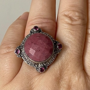 May include: A silver ring with a large, round, pink gemstone surrounded by smaller pink gemstones. The ring is set in a square-shaped design.