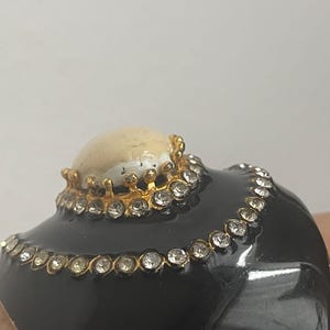 Puede incluir: Una pulsera brazalete ancha de color negro con una piedra cabuj&oacute;n central lisa de color crema engastada en un bisel dorado en forma de corona. La pulsera est&aacute; decorada con dos filas conc&eacute;ntricas de brillantes transparentes en metal dorado.