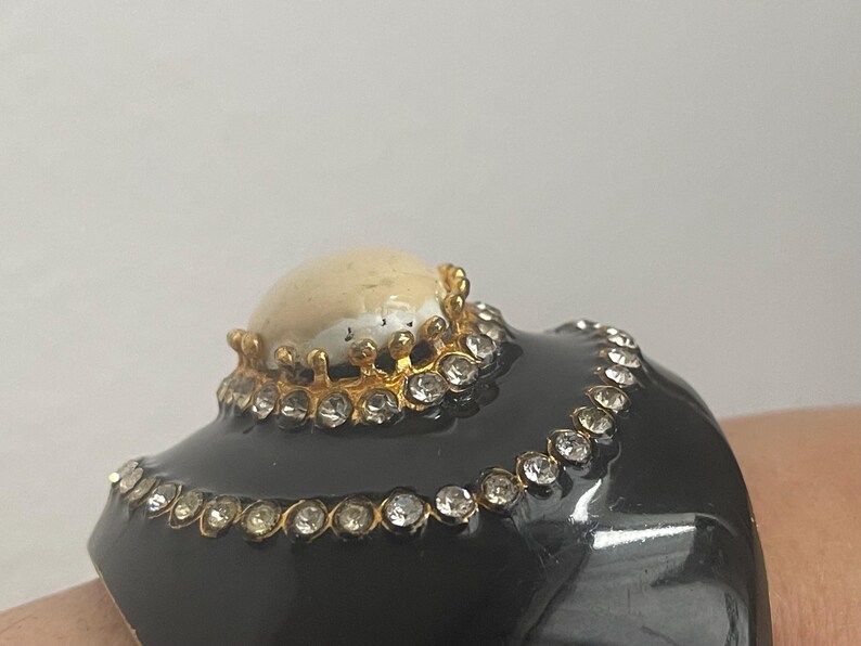 Puede incluir: Una pulsera brazalete ancha de color negro con una piedra cabuj&oacute;n central lisa de color crema engastada en un bisel dorado en forma de corona. La pulsera est&aacute; decorada con dos filas conc&eacute;ntricas de brillantes transparentes en metal dorado.
