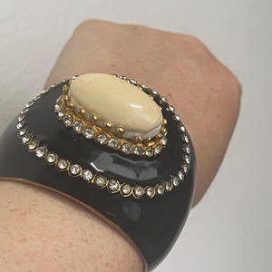 Puede incluir: Una pulsera brazalete ancha de color negro con una piedra cabuj&oacute;n ovalada de color crema engastada en un marco de metal dorado. La pulsera est&aacute; decorada con dos filas conc&eacute;ntricas de brillantes transparentes que rodean la piedra central.