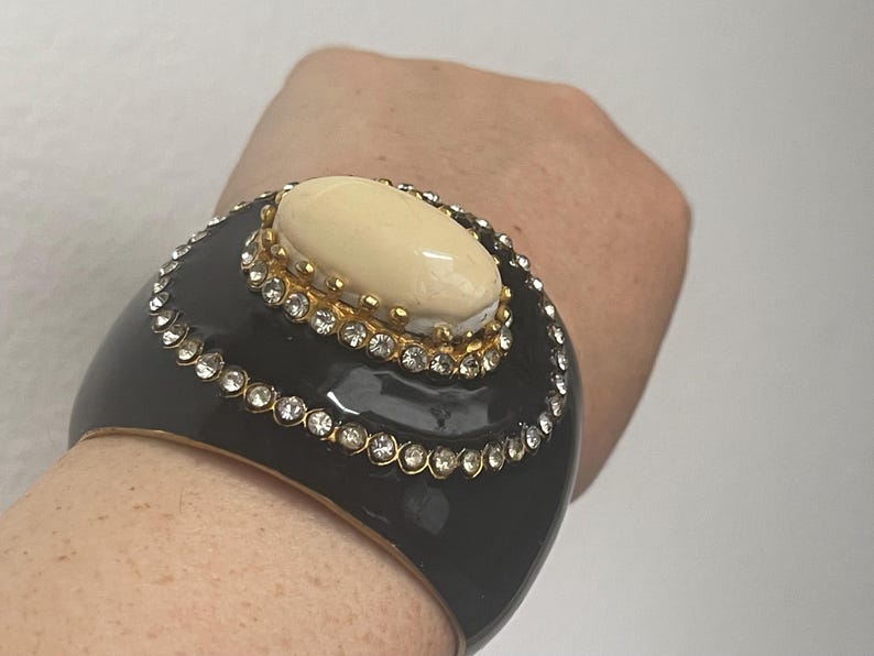 Puede incluir: Una pulsera brazalete ancha de color negro con una piedra cabuj&oacute;n ovalada de color crema engastada en un marco de metal dorado. La pulsera est&aacute; decorada con dos filas conc&eacute;ntricas de brillantes transparentes que rodean la piedra central.