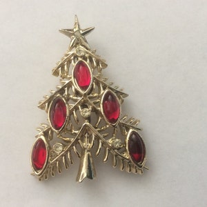 May include: Gold-tone Christmas tree brooch with red gemstone accents. The brooch features a star at the top and five oval-shaped red gems. Small clear crystals are also set in the design. A festive holiday accessory.