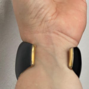 Puede incluir: Un brazalete ancho tipo pu&ntilde;o con acabado en negro mate y detalles de metal en tono dorado pulido en los extremos abiertos. El brazalete tiene un dise&ntilde;o curvo y suave que rodea la mu&ntilde;eca, destacando el contraste entre la superficie oscura y los bordes met&aacute;licos.