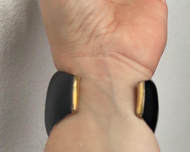 Puede incluir: Un brazalete ancho tipo pu&ntilde;o con acabado en negro mate y detalles de metal en tono dorado pulido en los extremos abiertos. El brazalete tiene un dise&ntilde;o curvo y suave que rodea la mu&ntilde;eca, destacando el contraste entre la superficie oscura y los bordes met&aacute;licos.