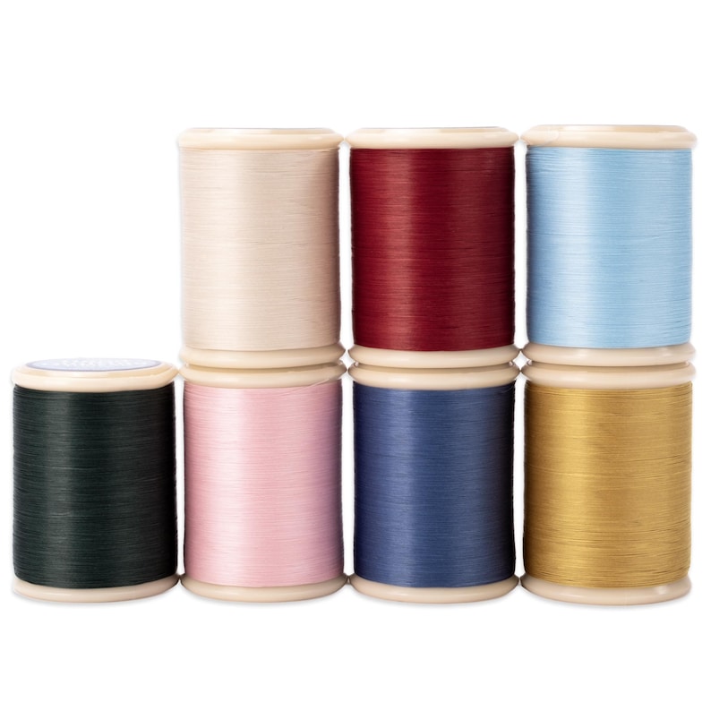 May include: A collection of eight spools of thread in various colors, including cream, burgundy, light blue, dark green, pink, navy blue, and gold. The spools are arranged in two rows, with the top row containing three spools and the bottom row containing five.