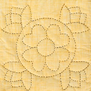 May include: A square, quilted fabric with a yellow background and a floral design. The design is created with black, dashed stitching, forming a large central flower and surrounding petal shapes. The fabric has a subtle, textured appearance.